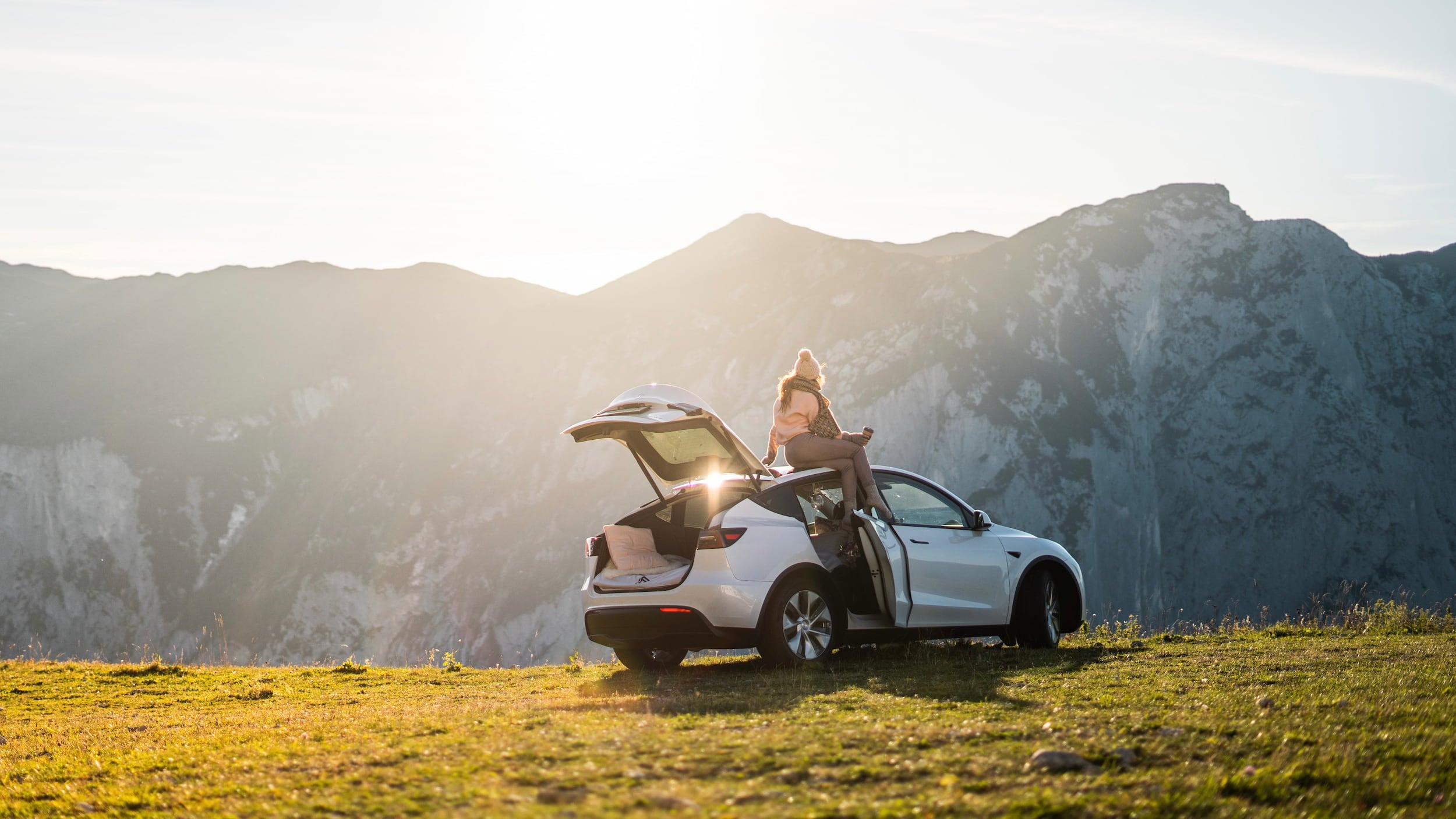 Woman sitting on top of a white Tesla Model Y parked on a mountain ridge during a car camping road trip